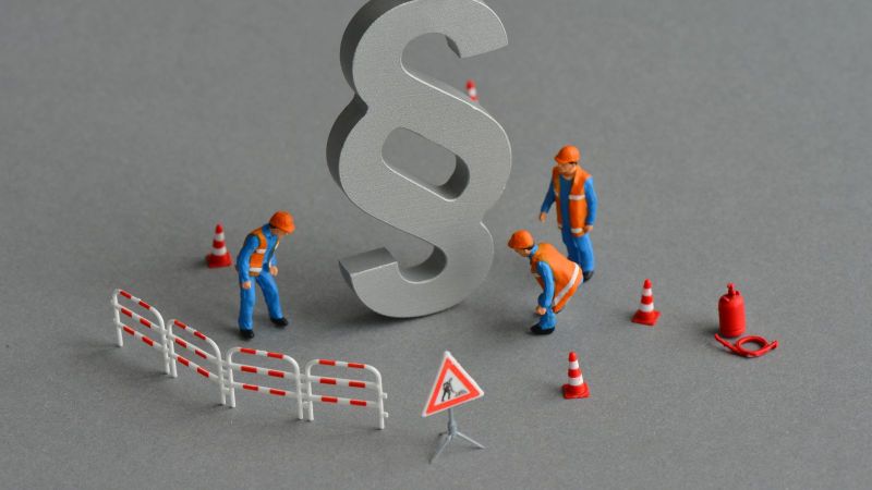 Das Bild zeigt eine gebaute Szene mit Spielzeugfiguren, die eine Baustelle um ein Paragrafenzeichen aufbauen.