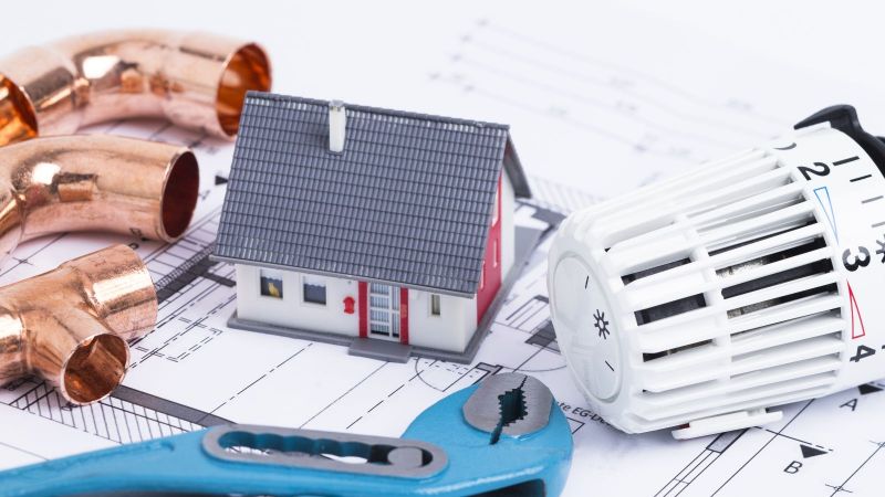 Das Bild zeigt einen Heizkopf, eine Rohrzange, ein Heizrohr und eine Mini-Modell eines Einfamilienhauses, die auf einem Bauplan platztiert wurden. 