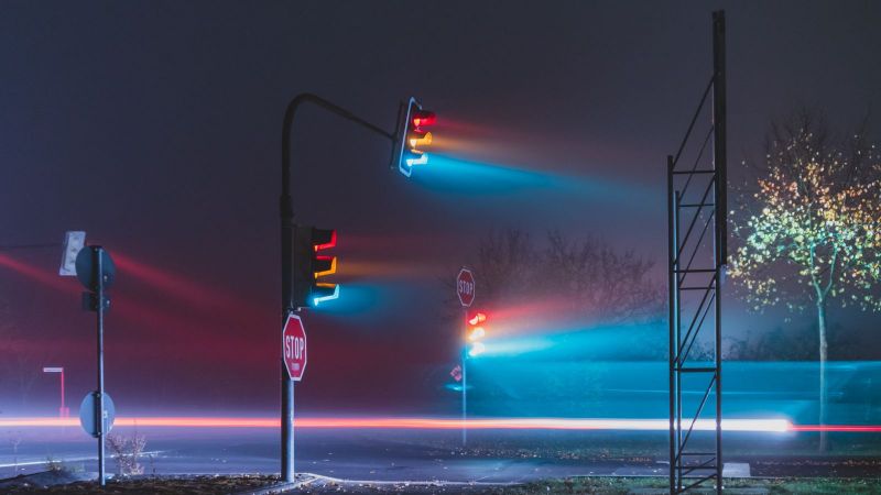 Das Foto zeigt eine leere Straße im Nebel, auf der drei Ampeln stehen, deren drei Lichter grün, gelb und rot allesamt leuchten. 