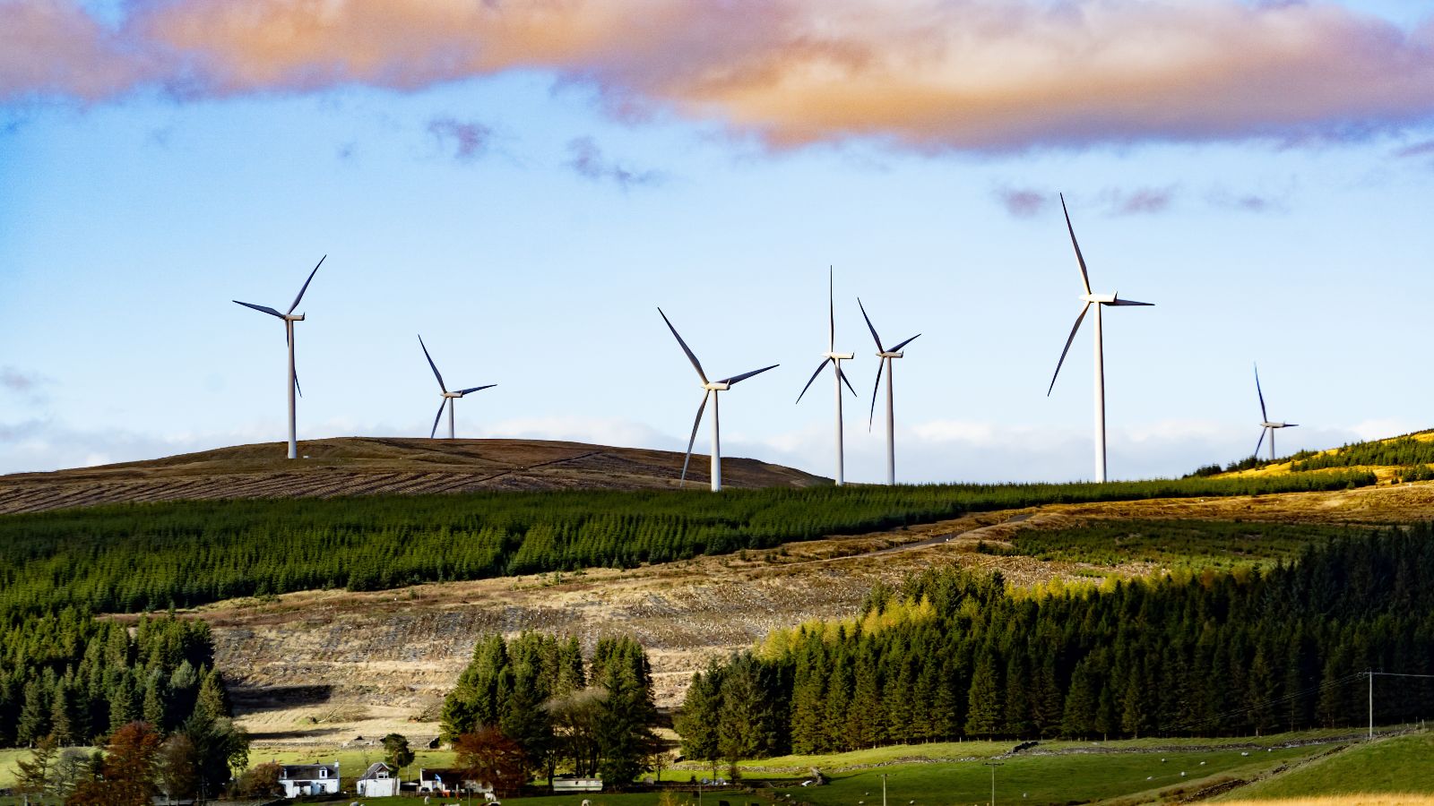 Das Bild zeigt drei Windräder auf einem Hügel einer ländlichen Region, in der sich auch ein Wohngebiet befindet. 