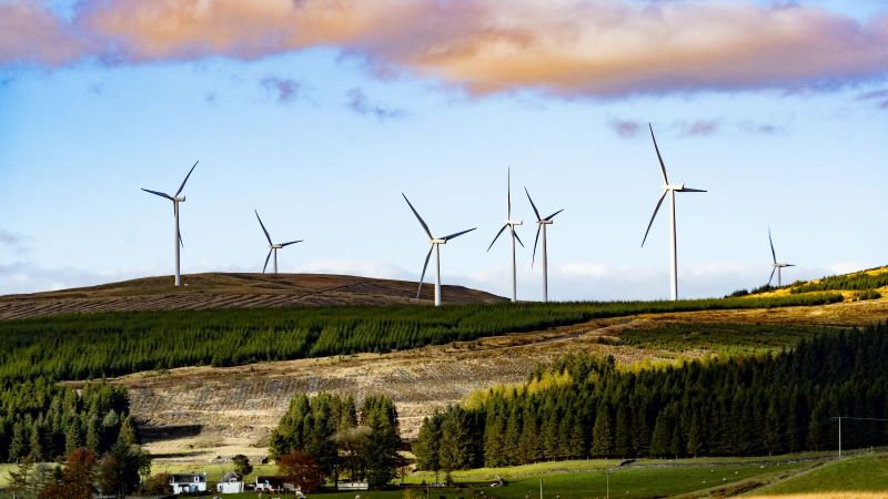 Das Bild zeigt drei Windräder auf einem Hügel einer ländlichen Region, in der sich auch ein Wohngebiet befindet. 