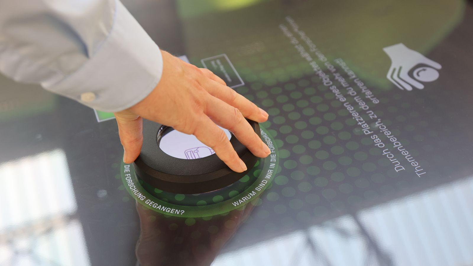 Das Bild zeigt eine Hand, die ein interaktives Element über einen mit Touchscreen ausgestatteten Tisch führt.