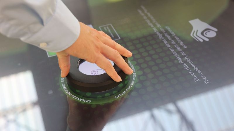Das Bild zeigt eine Hand, die ein interaktives Element über einen mit Touchscreen ausgestatteten Tisch führt.