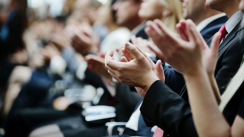 Das Bild zeigt sitzende Reihen von Menschen in formeller Kleidung, die applaudieren.