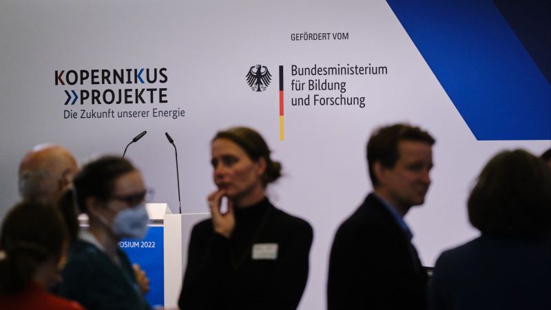 Das Bild zeigt angedeutet mehrere Symposiums-Besuchende im Diskurs, im Hintergrund deutlich das Kopernikus-Logo und das BMBF-Förderlogo.