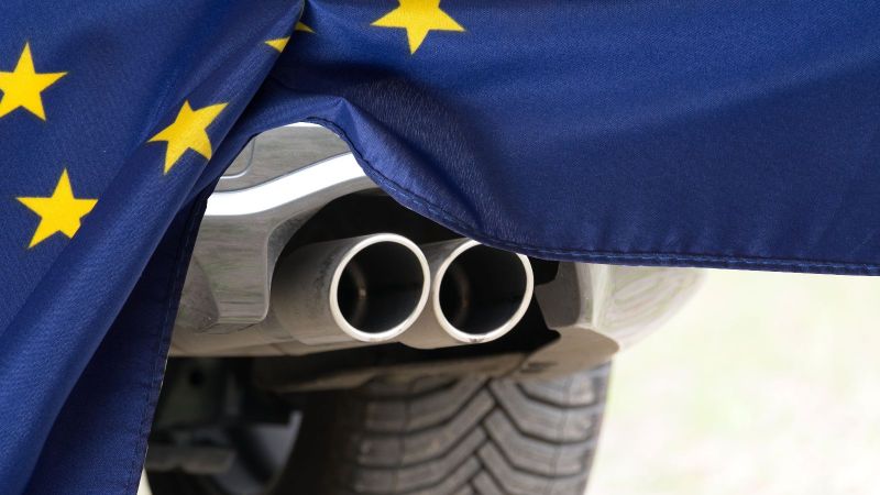 Das Bild zeigt den Auspuff eines motorisierten Fahrzeugs, der halb hinter einer Europafahne verschwindet.