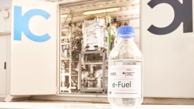 Das Bild zeigt eine durchsichtige Flasche mit der Aufschrift E-Fuel vor einer im Hintergrund unscharf erkennbaren Apparatur