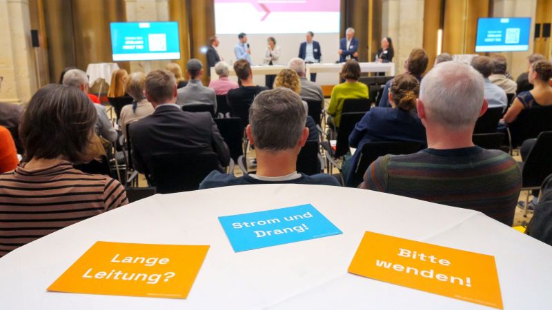 Das Foto zeigt Ariadne-Botschaften auf Postkarten, auf einem Tisch liegend. Dahinter sind Menschen in Stuhlreihen sitzend zu sehen, entfernt eine Podiumsdiskussion vor eine Ariadne-Hintergrund.