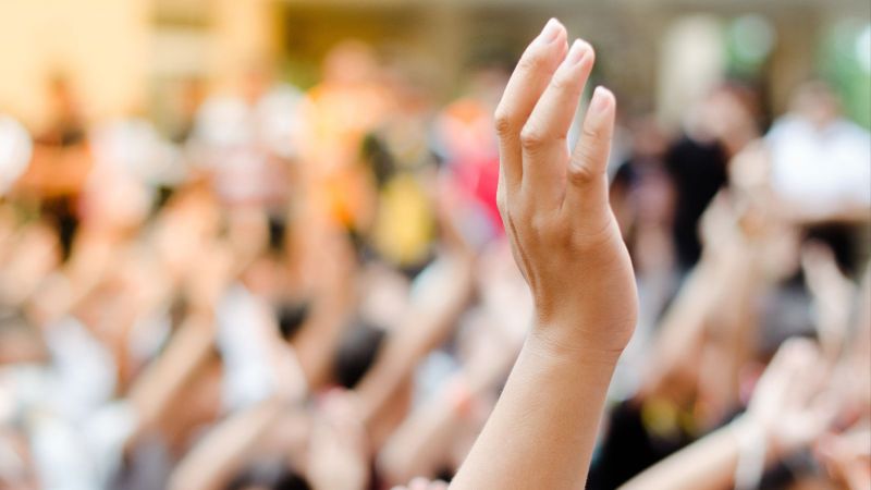 Das Bild zeigt eine unscharf angedeutete Menschenmenge und eine sich erhebende Hand, was symbolisch für Bürgerbeteiligung steht.