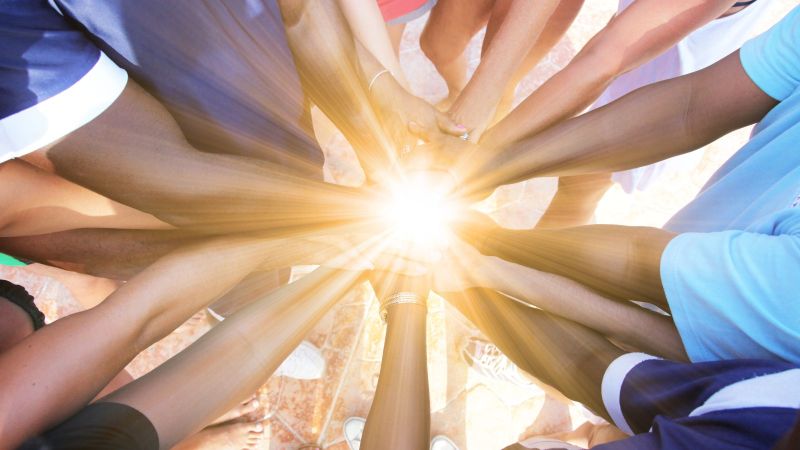 Das Bild zeigt, wie diverse Menschen sich in der Mitte die Hände reichen, während Sonnenstrahlen zu sehen sind.