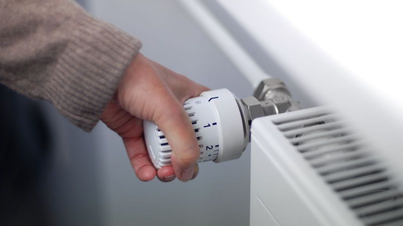 Das Bild zeigt eine Hand von einem mit einem dicken Pullover bekleideten Menschen, die das Heizungsthermostat runterdreht.