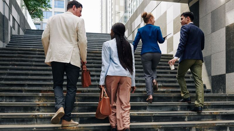 Das Bild zeigt zwei Personen, die von zwei weiteren Personen, die sie beraten und ihnen Fragen beantworten, eine Treppe hinauf begleitet werden.