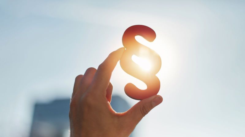 Das Bild zeigt eine Hand, die  ein Paragraphen-Zeichen ins Sonnenlicht hält.