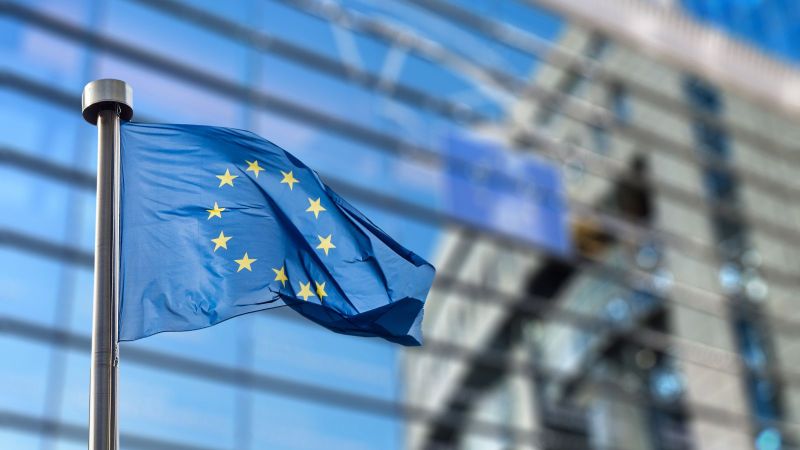 Das Bild zeigt eine Europaflagge, die vor der Spiegelung eines Gebäudes auf einem anderen Gebäude weht.