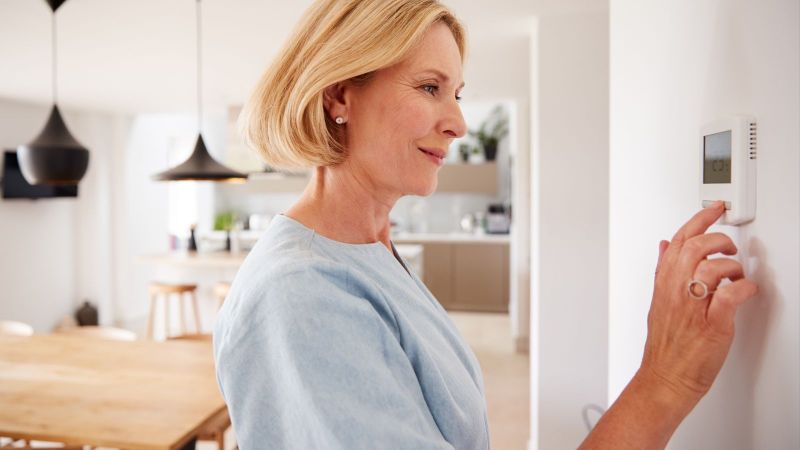 Das Bild zeigt eine Frau in einem Wohnraum, die vor einem digitalen Temperaturregler an der Wand steht und diesen bedient.