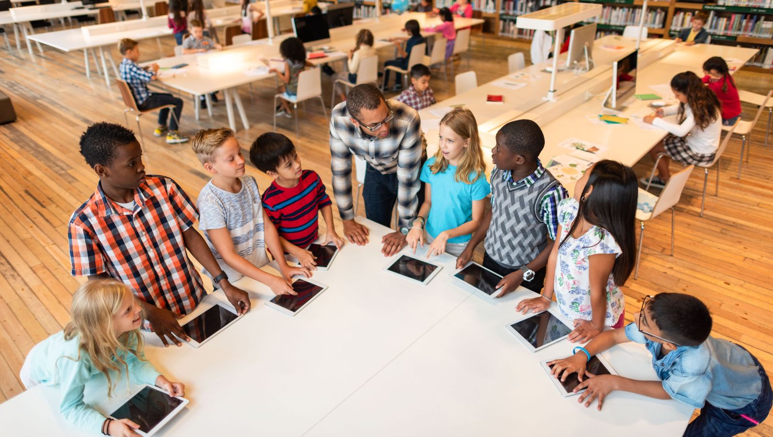 The picture shows a group of children sitting at a table with tablets, listening to an adult explaining something to them in a library setting.