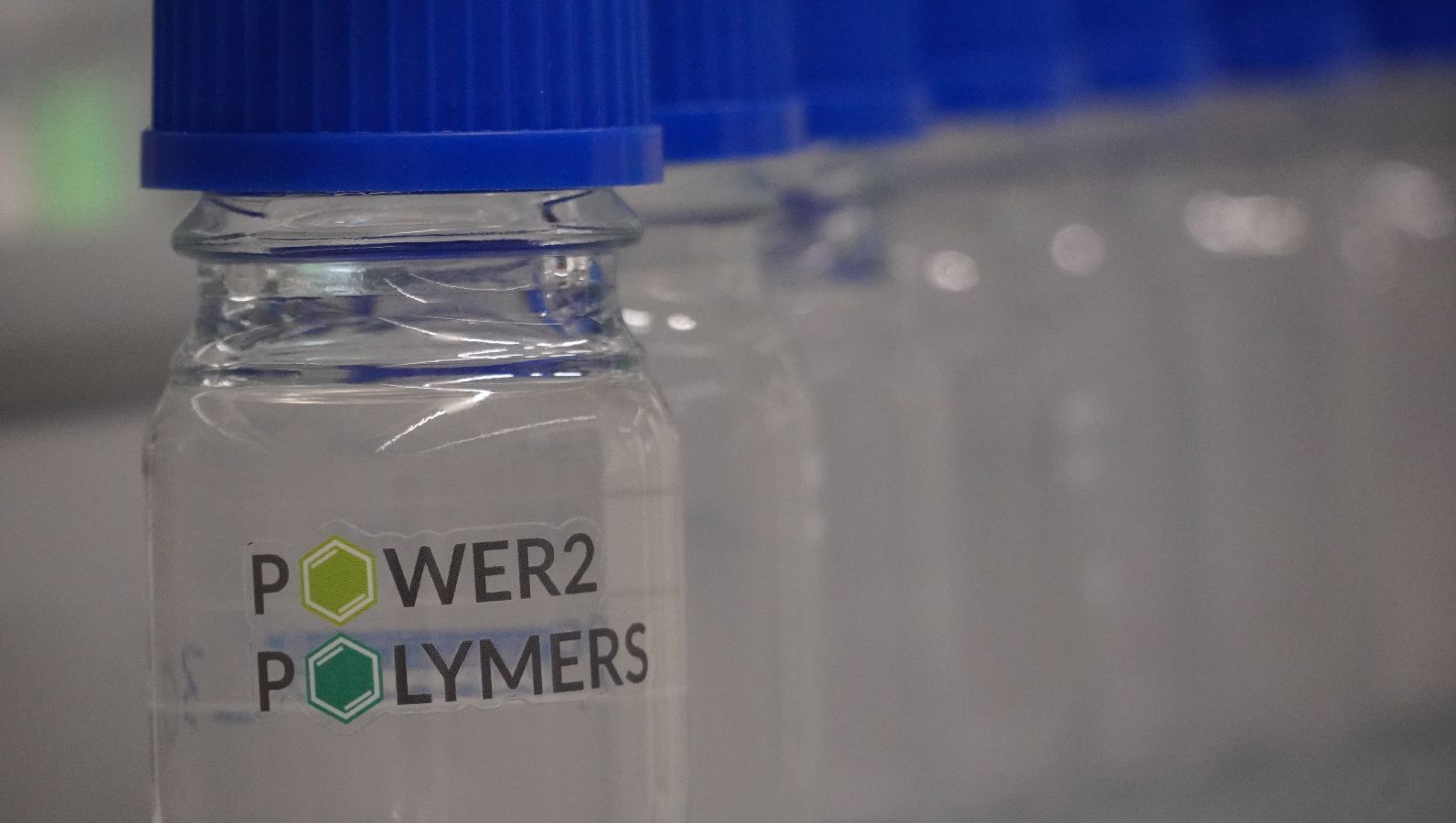 Das Bild zeigt mehrere Glasbehälter mit durchsichtigem flüssigen Inhalt und dem Power2Polymers-Logo als Aufdruck.