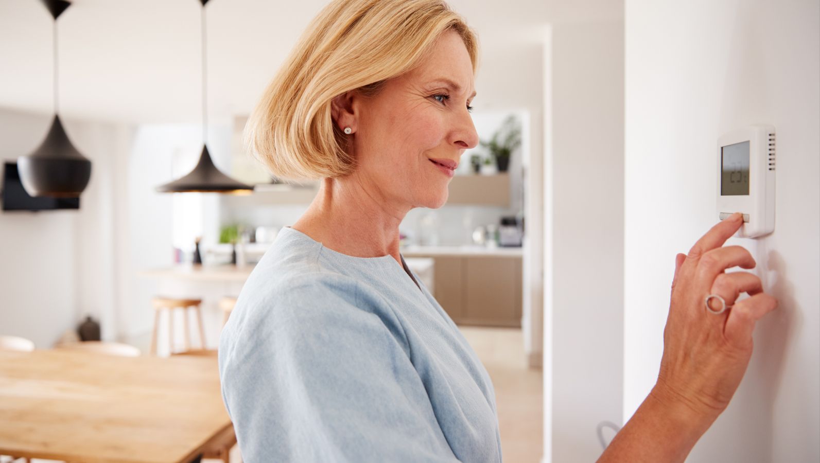 Das Bild zeigt eine Frau im Innenraum eines Hauses. Sie steht vor einer Wand und hat eine Hand an einem Klimaregler oder einem Smart Meter an der Wand.