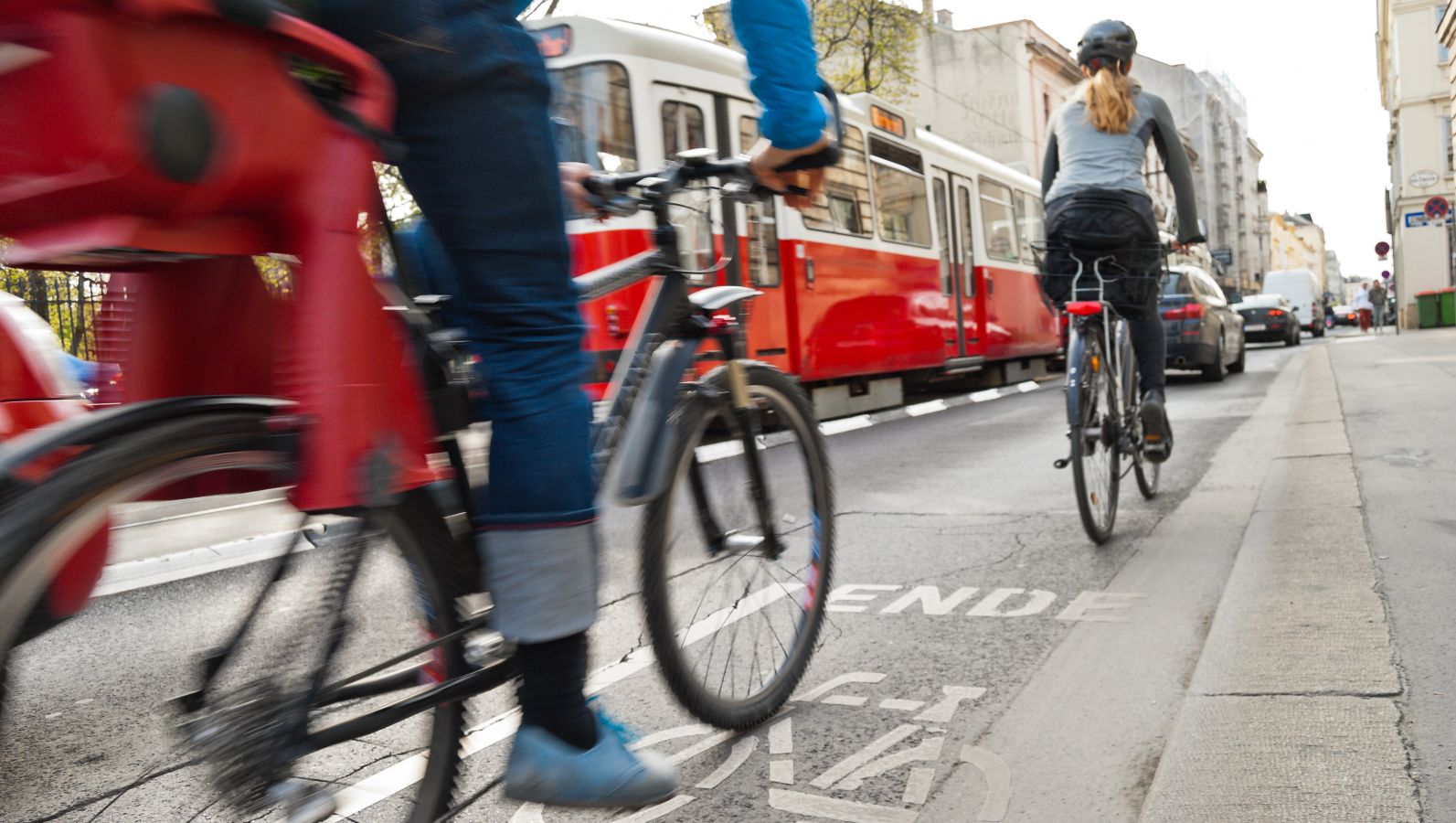 Das Bild zeigt Fahrradfahrende von hinten auf einem Fahrradstreifen neben einer städtischen stark befahrenen Straße mit motorisierten Verkehrsmitteln.