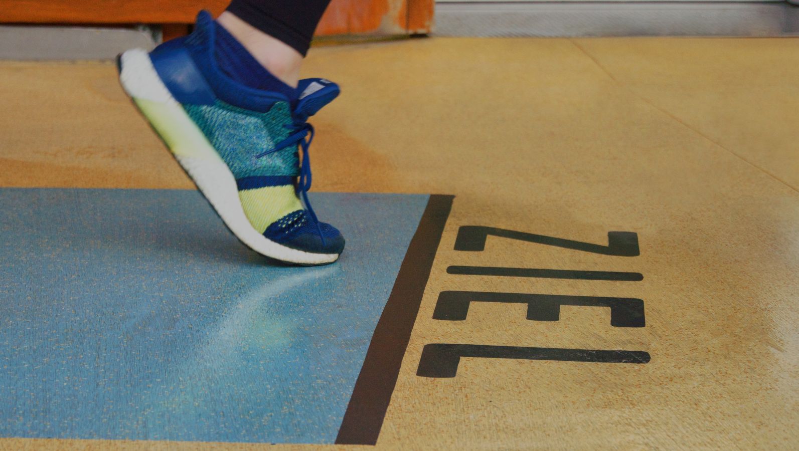 Zu sehen ist ein Fuß in einem Sportschuh von einer Person, die offenkundig in einer Laufbewegung ist; der Fuß befindet sich unmittelbar vor einer Linie, hinter der 