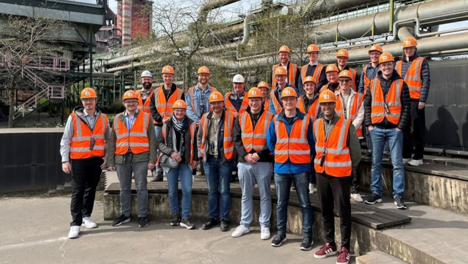 Zu sehen ist eine Gruppe junger Menschen in Schutzwesten und mit Schutzhelmen vor einem industriellen Hintergrund.