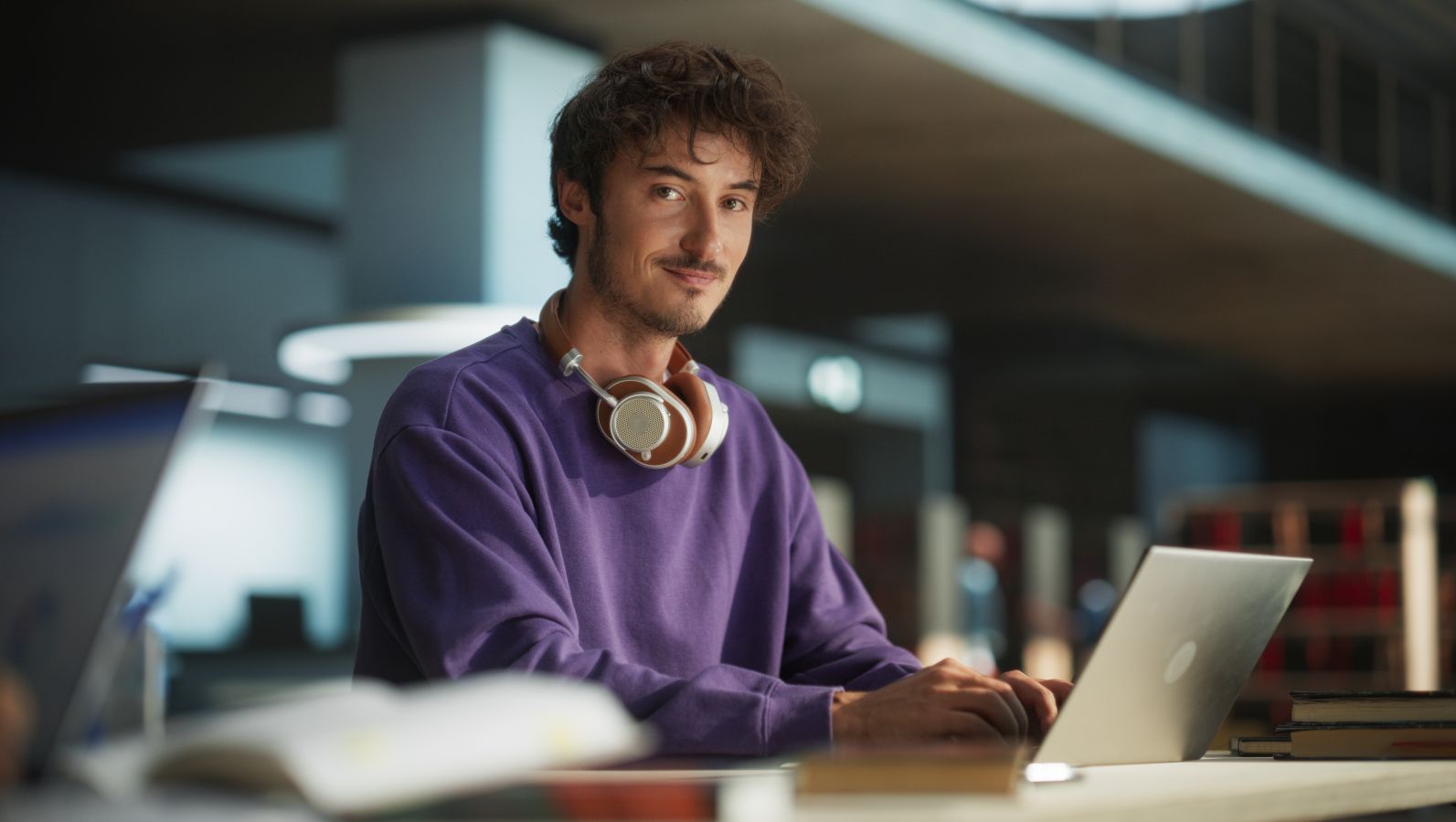Das Foto zeigt einen jungen Mann in einer Umgebung, die nach Bibliothek aussieht, lächlnd und mit Kopfhörern an einem Computer.