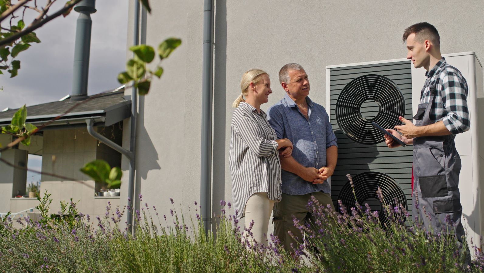 Das Bild zeigt einem Techniker, der einem Paar neben einem Haus und einer Wärmepumpe etwas erklärt.