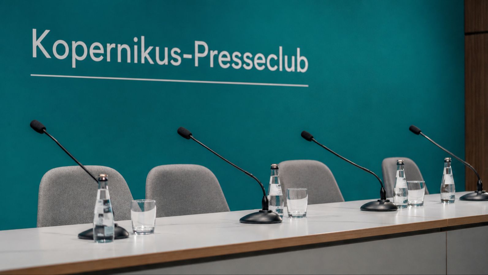 Bild von Tisch und Stühlen mit Mikrofonen für eine Pressekonferenz mit einem Hintergrund, auf dem 